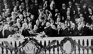 President Harding faces the Chief Justice with his hand raised to take the Oath of Office. A large crowd stands behind him.