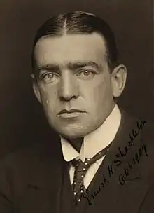 Man with hair centre-parted, wearing high white collar with tie, and a dark jacket. His facial expression is serious