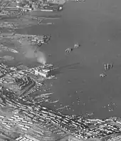 A black and white photograph of the San Francisco Bay shoreline at Hunters Point. The Hunters Point Power Plant is distinguished in the center of this cropped photograph as a large facility emitting smoke from a tall smokestack.