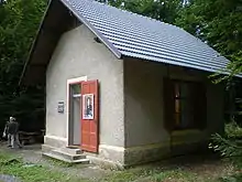 A small grey hut, surrounded by woods, with an open door to which is affixed a picture of the composer