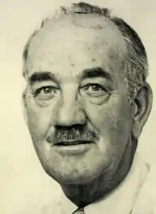 Black and white photo of a man with balding black and grey hair and a small moustache, wearing a white dress shirt and a necktie