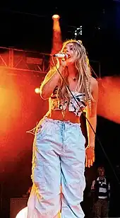 A blonde woman in a white and black patterned crop top and light blue jeans wearing a crocheted frog hat.