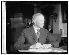 Black and white photograph of a seated, short-haired man looking off to the side and wearing a striped suit and tie.