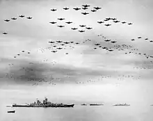 Black and white photo showing a large number of aircraft flying in formation over several World War II-era warships
