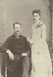 A black-and-white photograph of a couple. On the left is a seated man with a moustache weraing a dark suit. Standing on the right is a woman in a white dress, body facing left. Both face the camera.