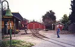 An elderly man standing next to railroad track in the foreground with an old-fashioned railroad depot building on the opposite side. A locomotive shed and water tower are located where the railroad track ends in the background.