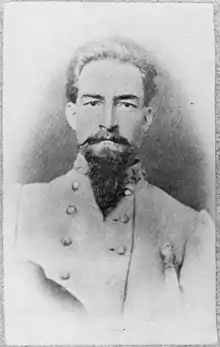 Photo shows a solemn man with a dark moustache and beard looking directly at the camera. He wears a gray military uniform with a star visible on the collar.